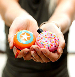 A Super hero orange bike bell and a multi-coloured dotted bike bell held in womens hands